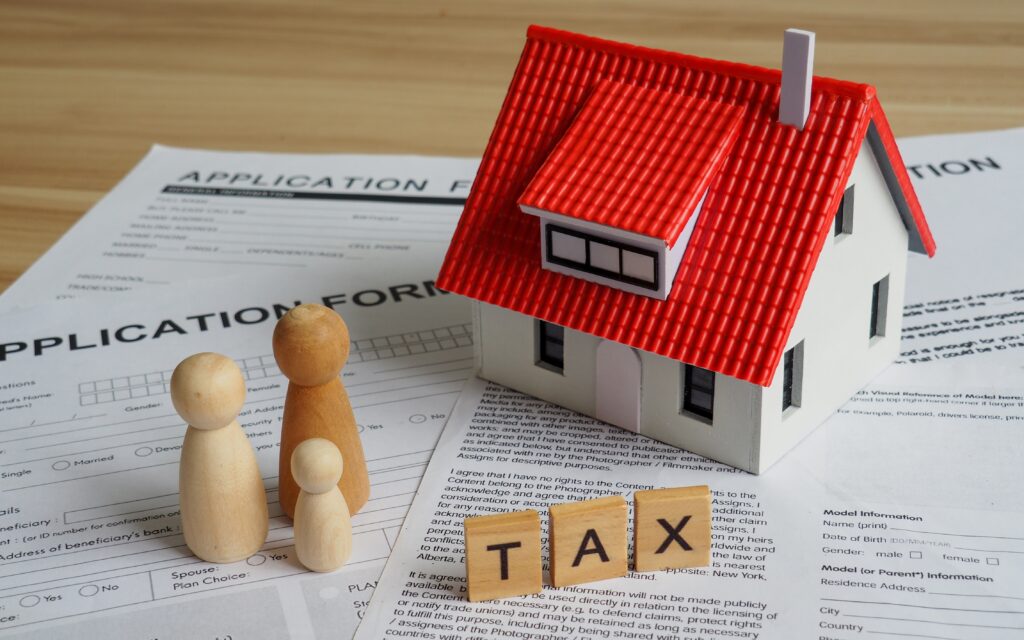 Miniature model house on a table with application papers, models of a mini family and tiles that spell out the word task. All together, they symbolize the Ontario Land Transfer Tax Rebate for First-Time Home Buyers